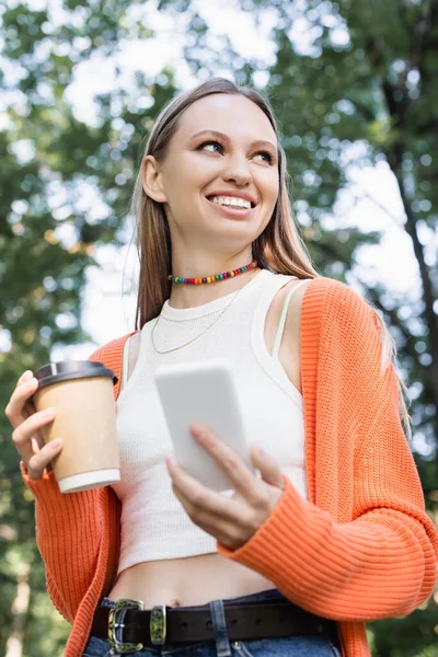 Vista de ángulo bajo de la mujer feliz sosteniendo taza de papel y teléfono inteligente - foto de stock