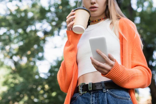 Низкий угол зрения обрезанной женщины с бумажной чашкой и смартфоном в зеленом парке — стоковое фото