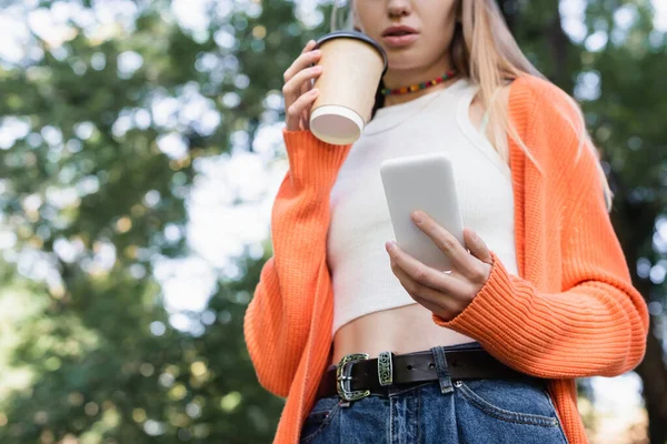 Vista de ángulo bajo de la mujer recortada sosteniendo taza de papel y teléfono inteligente en el parque verde - foto de stock