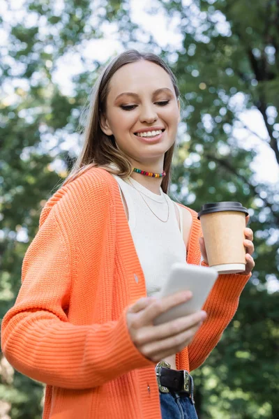 Low angle view of cheerful woman holding paper cup and using smartphone - foto de stock