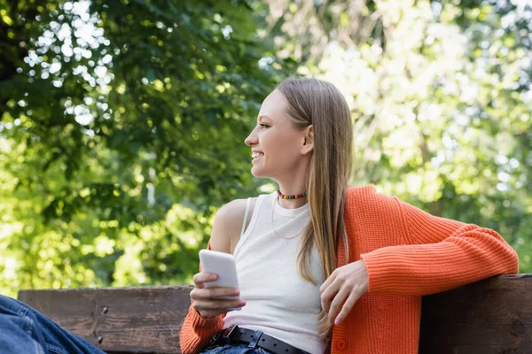 Femme heureuse tenant smartphone et assise sur un banc — Photo de stock