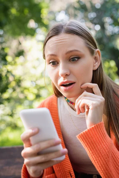 Surprised woman using blurred smartphone in green park — Stockfoto