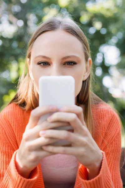 Жінка прикриває обличчя при використанні розмитого смартфона в зеленому парку — стокове фото
