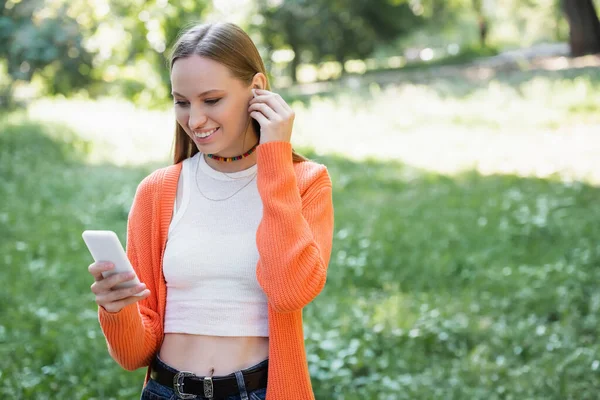 Cheerful woman wearing wireless earphone and using smartphone in park — Stock Photo