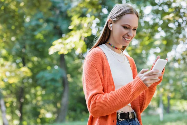 Cheerful woman in wireless earphone listening music and using smartphone in park — Stockfoto