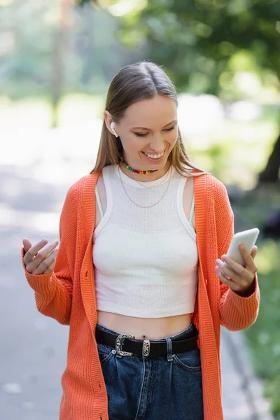 Positive woman in wireless earphone using mobile phone in green park — Fotografia de Stock