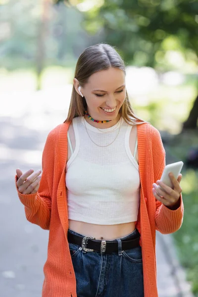 Positive woman in wireless earphone using mobile phone in green park — Stock Photo
