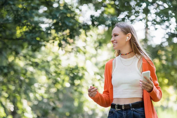 Joyful woman in wireless earphone holding smartphone in park — Stock Photo