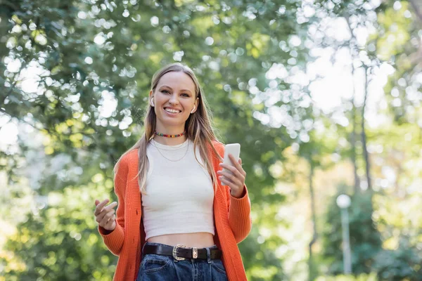 Donna sorridente in auricolari senza fili che tiene smartphone nel parco — Foto stock