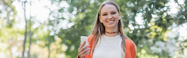 Задоволена жінка в помаранчевому кардигані та бездротових навушниках за допомогою смартфона в парку, банер — стокове фото
