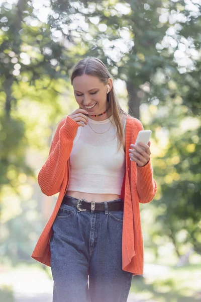 Cheerful woman in orange cardigan and wireless earphone using smartphone in park — Stockfoto