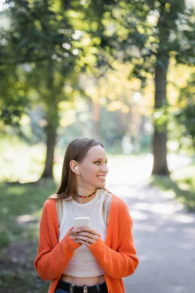 Pleased woman in orange cardigan and wireless earphone using smartphone in park — Stock Photo