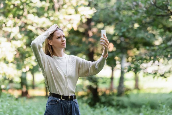 Worried young woman with smartphone searching mobile service in park — Stockfoto