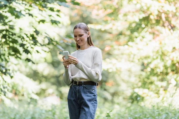 Весела молода жінка зі смартфоном, що дивиться в зелений парк — стокове фото