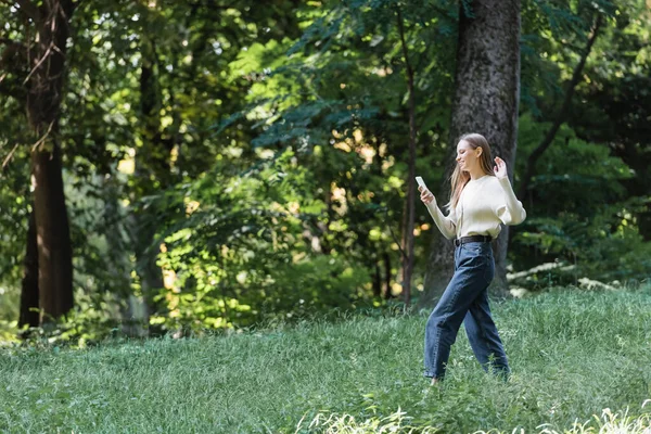 Happy young woman waving hand during video call on smartphone while walking in park — Stock Photo