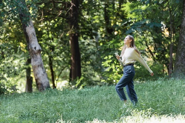 Happy young woman with smartphone walking in green park — Foto stock