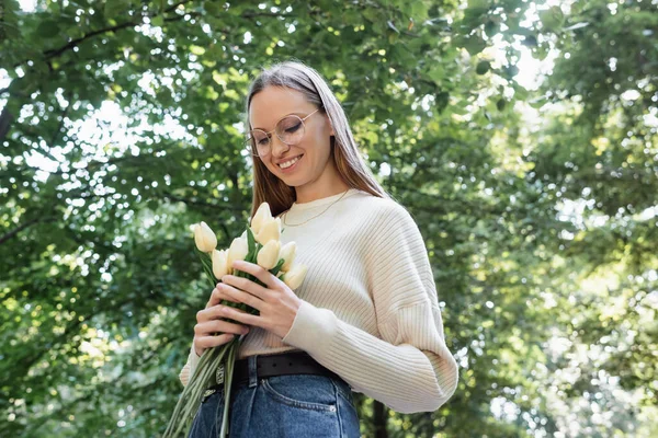 Low angle view of happy woman in glasses holding tulips in green park — Fotografia de Stock