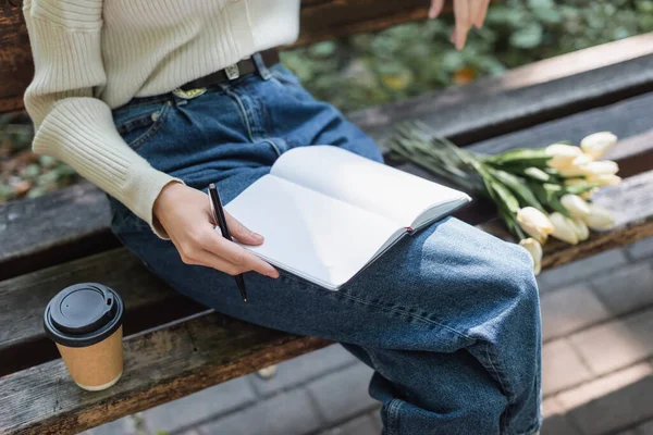 Vista ritagliata di donna in jeans che tiene notebook vicino a mazzo di tulipani e tazza di carta su panchina — Foto stock
