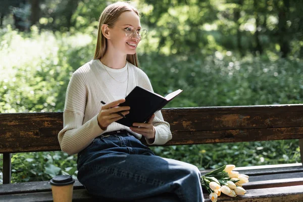 Fröhliche Frau in Jeans hält Notizbuch neben Tulpenstrauß und Pappbecher auf Bank — Stockfoto