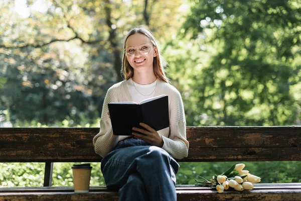 Cheerful woman in glasses holding notebook near bouquet of tulips and paper cup on bench — Stock Photo