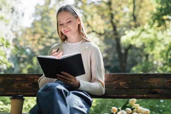 Cheerful woman looking at notebook near bouquet of tulips and paper cup on bench — Fotografia de Stock