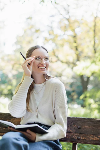 Felice donna che regola gli occhiali mentre tiene il notebook nel parco verde — Foto stock