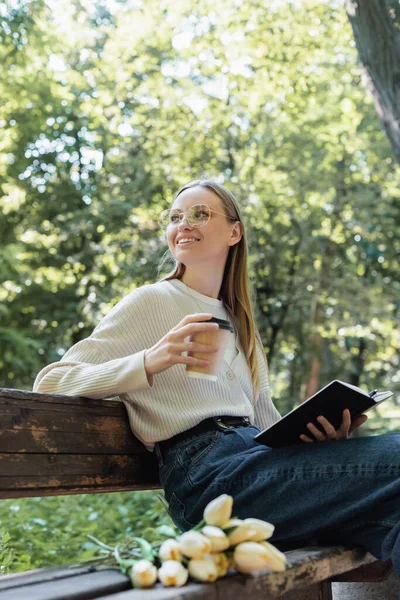 Vista basso angolo di donna sorridente che tiene tazza di carta e notebook mentre si siede sulla panchina — Foto stock