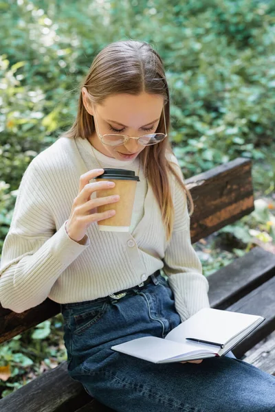 Woman holding paper cup and looking at notebook while sitting on bench — Foto stock