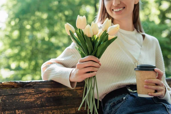 Cropped view of happy woman holding bouquet of blooming tulips and paper cup in park — Stock Photo