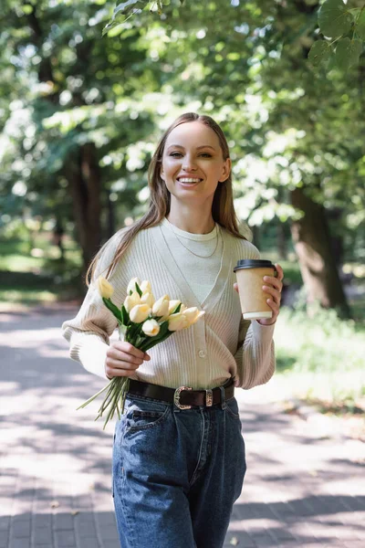 Cheerful woman in glasses and jeans walking with tulips and paper cup in park — Fotografia de Stock