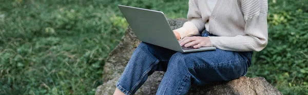 Cropped view of woman using laptop while sitting on stone, banner — Stockfoto