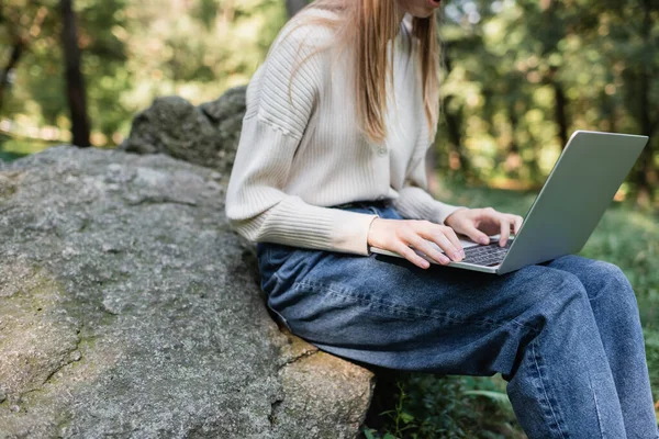 Cropped view of woman using laptop while sitting on stone - foto de stock