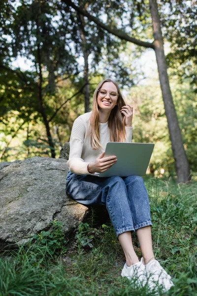 Happy woman in glasses using laptop while sitting on stone in park - foto de stock