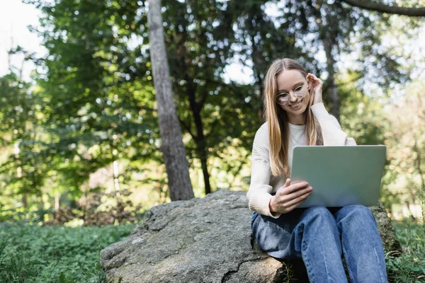 Low angle view of happy woman in glasses adjusting hair and looking at laptop in park — Stock Photo
