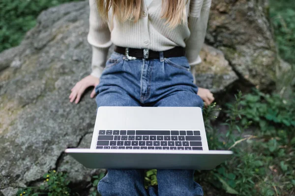 Vista ritagliata di freelance in jeans utilizzando il computer portatile mentre seduto sulla pietra — Foto stock