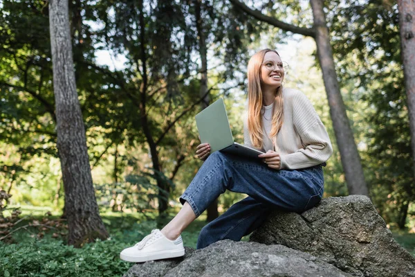 Donna felice in bicchieri che tiene il computer portatile mentre si siede sulla pietra nel parco — Foto stock