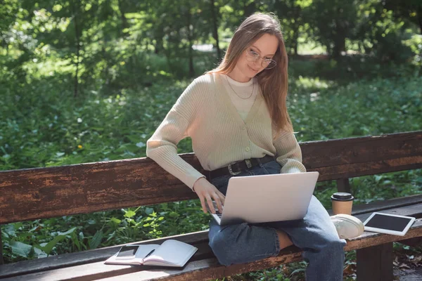 Heureux pigiste dans des lunettes à l'aide de gadgets pour ordinateur portable soigné et café pour aller sur le banc — Photo de stock