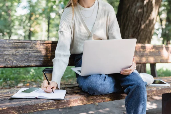 Vista ritagliata della donna che usa il computer portatile mentre scrive nel notebook vicino ai gadget sulla panchina — Foto stock