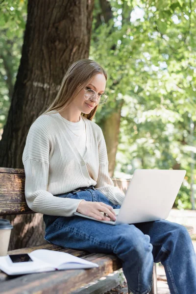 Woman in glasses using laptop and sitting on bench in green park — Stock Photo
