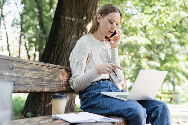 Woman in glasses talking on smartphone and using laptop while sitting on bench in green park — Stockfoto