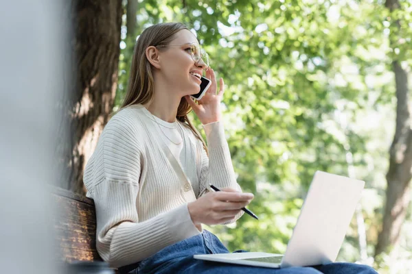 Happy woman in glasses talking on smartphone and using laptop while sitting on bench — Stockfoto