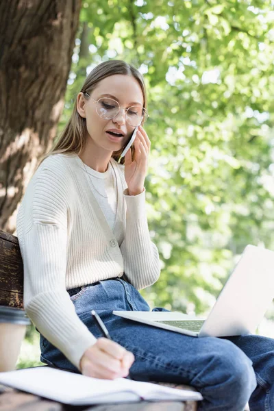 Mulher de óculos falando no smartphone e escrevendo no notebook enquanto sentado com laptop no banco — Fotografia de Stock