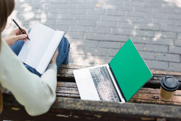 Top view of woman writing in notebook near laptop with green screen and paper cup — Foto stock