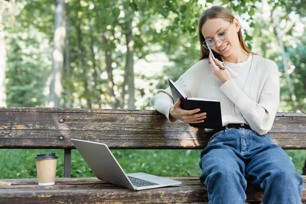 Fröhliche Frau mit Brille, die auf dem Smartphone spricht, während sie neben dem Laptop auf der Bank sitzt — Stockfoto