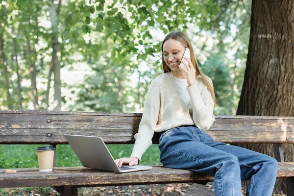 Cheerful woman in glasses talking on smartphone while using laptop on bench - foto de stock