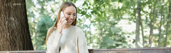 Cheerful woman in glasses talking on smartphone in green park, banner — стоковое фото