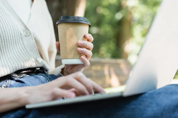 Cropped view of woman using laptop while holding paper cup — стоковое фото
