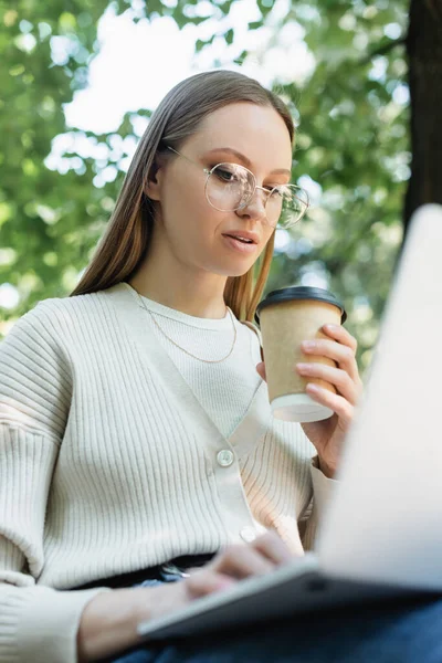 Femme dans les lunettes à l'aide d'un ordinateur portable tout en tenant tasse en papier dans le parc vert — Photo de stock