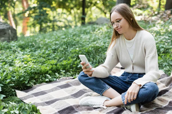 Усміхнена жінка в окулярах, використовуючи смартфон, сидячи з схрещеними ногами на картатому плед — стокове фото