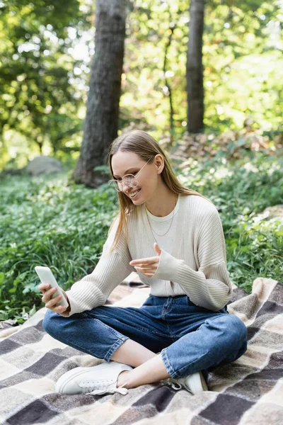 Улыбающаяся женщина в очках с помощью мобильного телефона, сидя со скрещенными ногами на клетчатой клетке — стоковое фото